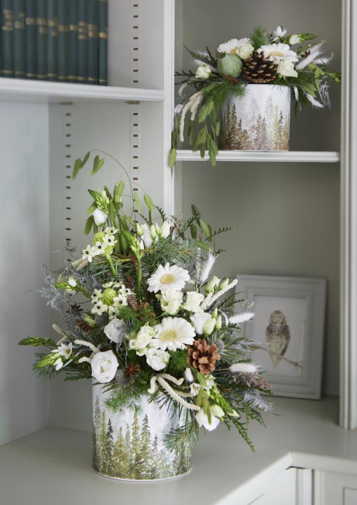 A white ceramic pot with a stunning forest fir tree pattern sits on a white shelf. Inside, there is a full easy Christmas floral arrangement featuring white roses, greenery, pine cones and more.
