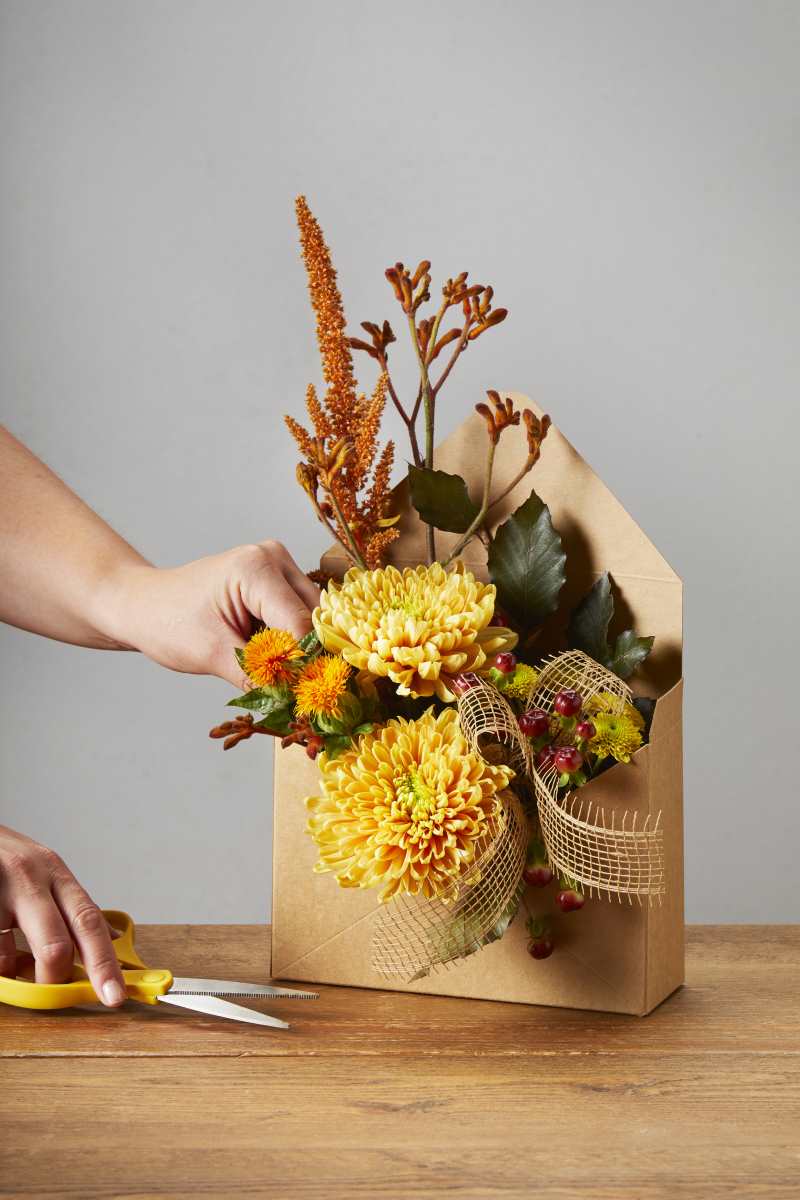Making an autumnal flower arrangement with a brown cardboard-style envelope box. There are yellow and burnt orange flowers, green foliage and a brown woven ribbon.