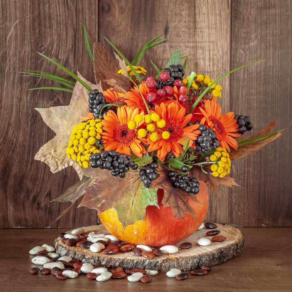 Halloween pumpkin flower arrangement with orange and yellow flowers, black and red berries and dried autumnal leaves. 