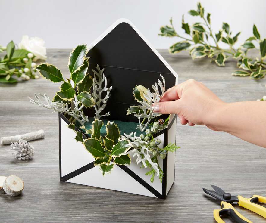 A black and white envelope box assembled and standing up right with the envelope flap opened. Somebody is placing green foliage inside. The envelope box is on a grey table. There are various floral crafting supplies and flowers on the table too.