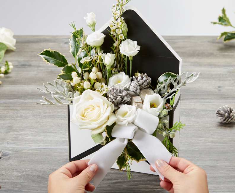 A black and white envelope box assembled and standing up right with the envelope flap opened. There is a floral foam arrangement inside made up of green foliage and various white flowers, including white roses. A velour ribbon is being tied to the front in a bow. The envelope box is on a grey table. There are various floral crafting supplies and flowers on the table too.