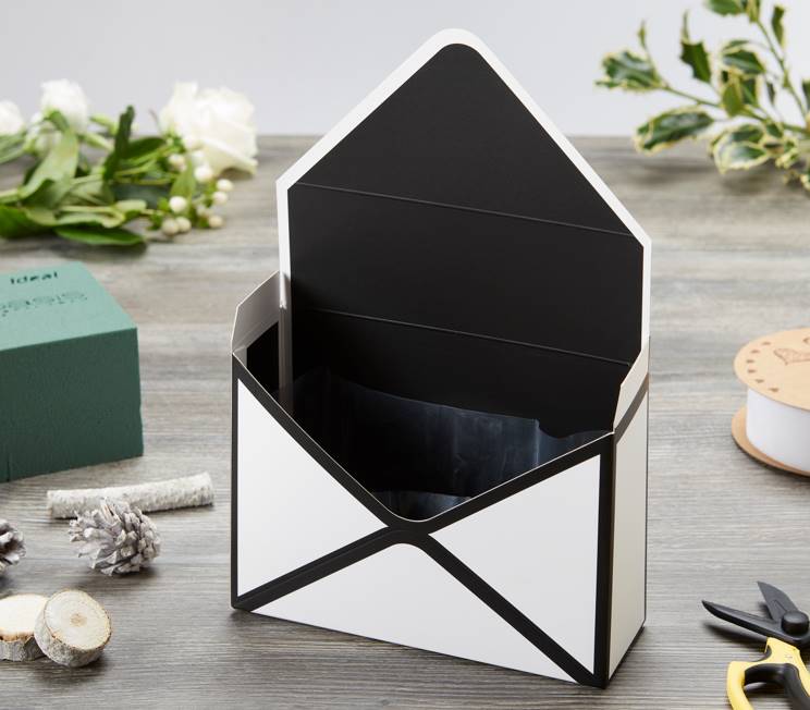 A black and white envelope box assembled and standing up right with the envelope flap opened. The envelope box is on a grey table. There are various floral crafting supplies and flowers on the table too.