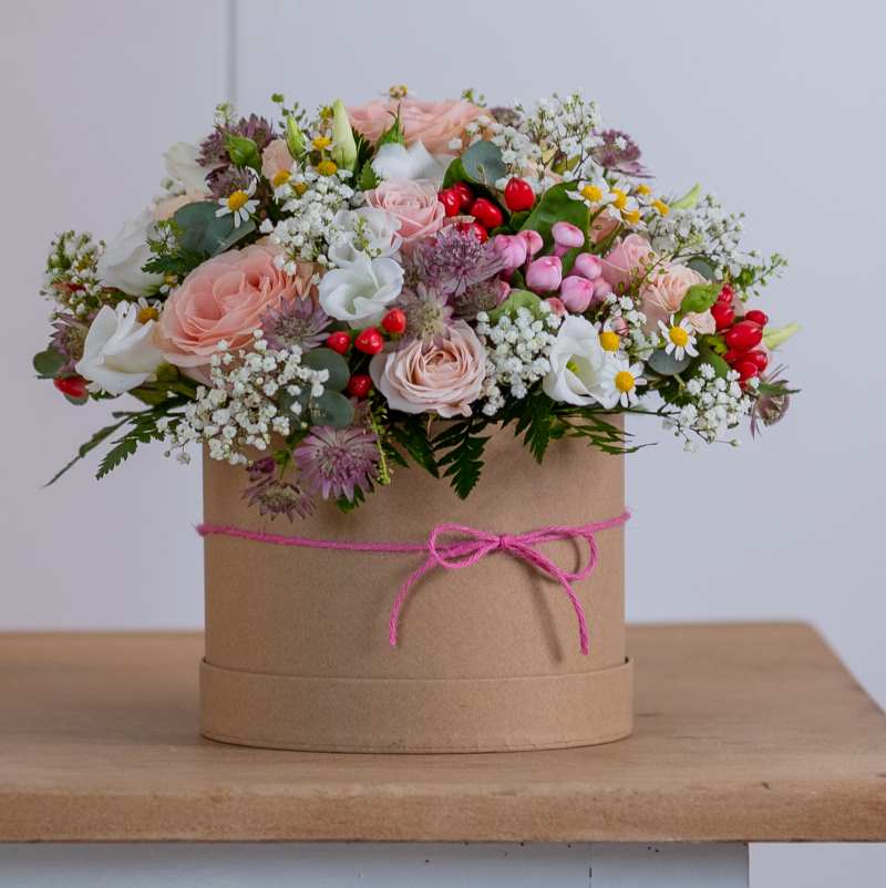 Brown natural-style hat box with a pink natural cord tied around and into a bow. A full floral arrangement is inside with light pink roses, gypsophila, red berries and green foliage.