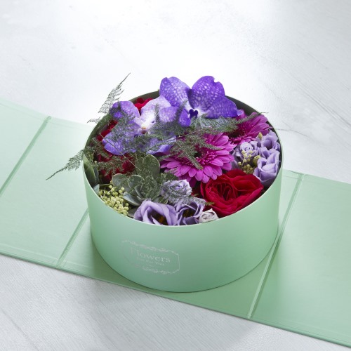 Mint green cylinder box with a beautiful floral arrangement featuring violets, red flowers, purple flowers and green foliage.