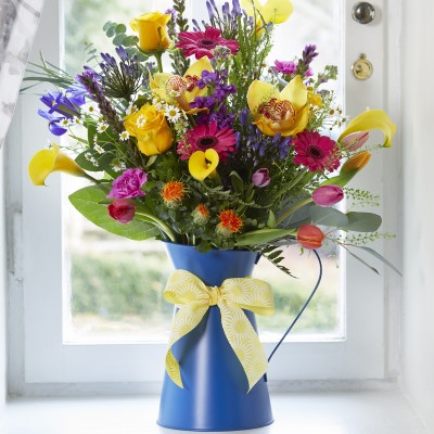 Blue Elijah jug with a yellow and white floral ribbon tied around it. The jug features a simple floral arrangement with a collection of different flowers in different colours, including yellow roses and pink tulips. There is also a good amount of foliage included.