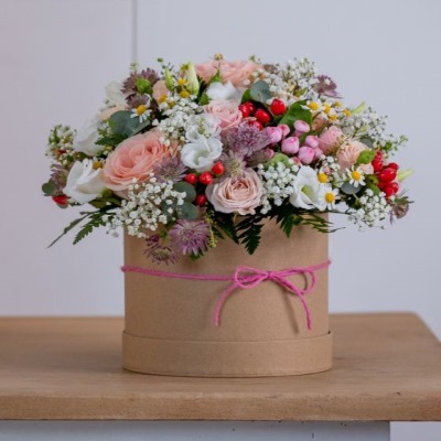 Simple floral arrangement using a brown craft hat box which has a pink ribbon tied around it. The flowers are a mix of white, pale pink, red and pale purple, and there is a good amount of green foliage.